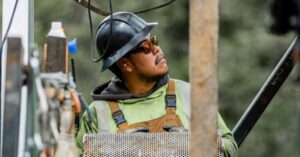 Construction worker wearing a hard hat and safety glasses, looking up at equipment outdoors with a metal mesh panel in front of them.